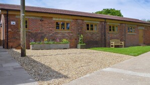 A brick building with a gravel driveway, a bench, and flower beds. - Woodlands Dairy Cottage (Adversane, near Billingshurst)