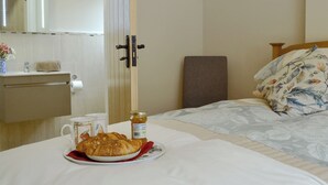 A hotel room with a bed, a bedside table with a plate of croissants and honey, a mirror, a wooden door, and a framed picture on the wall. - Woodlands Dairy Cottage (Adversane, near Billingshurst)
