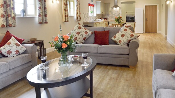 A spacious living room with a grey sofa, a coffee table, and a vase of flowers. There is a kitchen in the background with a dining area. - Woodlands Dairy Cottage (Adversane, near Billingshurst)