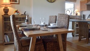 Un comedor con una mesa de madera dispuesta para cuatro personas, rodeada de sillas tejidas. Se ve un reloj de pared grande y un cuadro enmarcado.