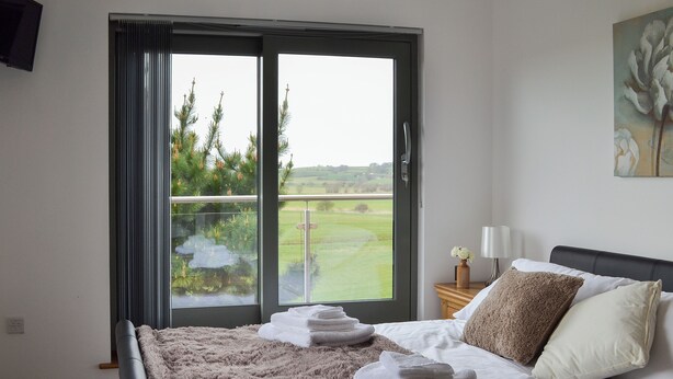 A bedroom with a bed, a nightstand, a lamp, and a view of greenery through a window. - 4 bedroom accommodation in Winchelsea, near Rye (Winchelsea, near Rye)