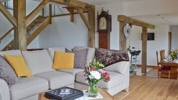 Un salon avec un canapé gris, des poutres en bois, une table à manger avec des chaises et une grande horloge murale.