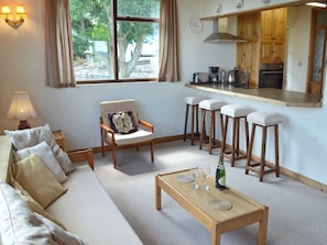Una sala de estar con un sofá, una mesa de centro de madera, una silla con cojín y una zona de cocina con taburetes.