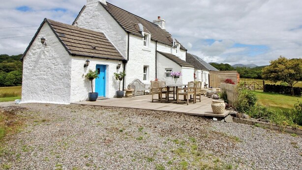 A white house with a blue door, a patio with wooden furniture, and a gravel driveway.
