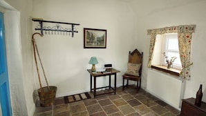 A room with a stone floor, a wooden chair, a table with a lamp, a picture on the wall, and a window with curtains.