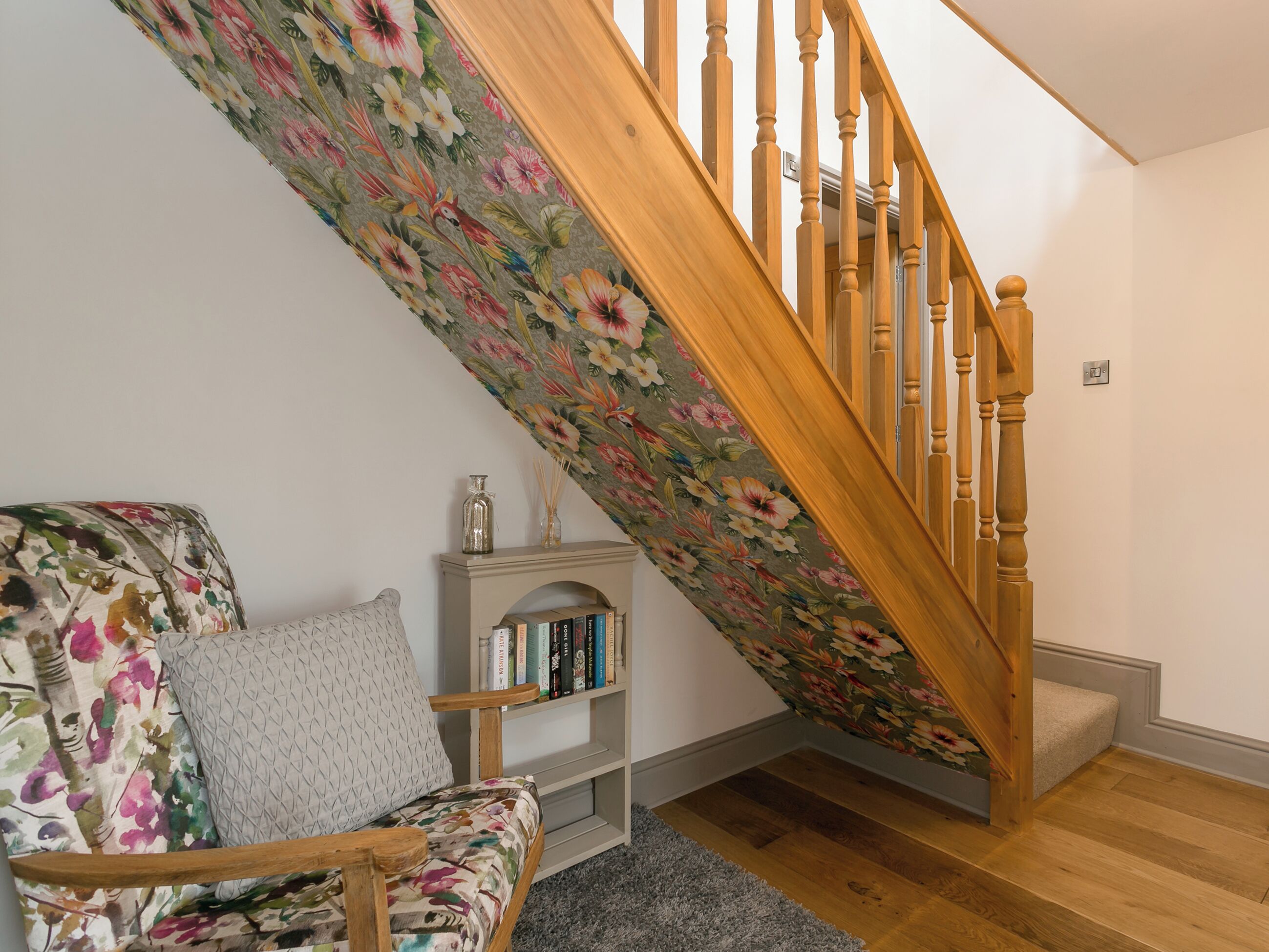 Een trap met houten spijlen, een wand met bloemenmotief, een boekenkast vol boeken en een gestreept fauteuil.