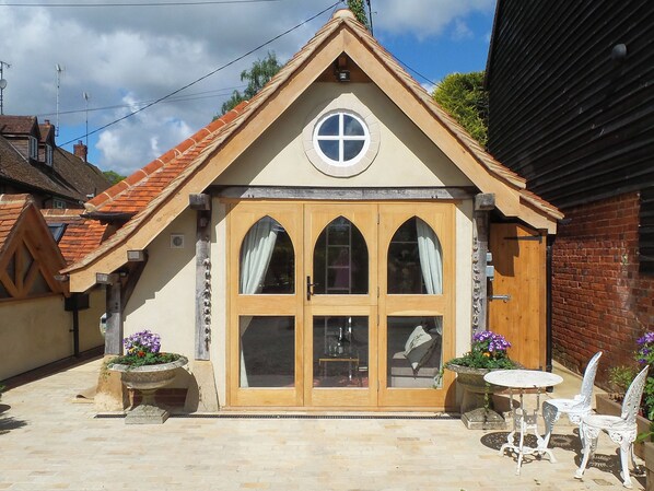 A building with a gabled roof, a central circular window, and a set of wooden double doors. There are two outdoor tables and chairs, and flower planters are visible.