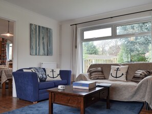 Una sala de estar con un sillón azul, un sofá color crema, una mesa de centro de madera y vistas al exterior a través de amplios ventanales.