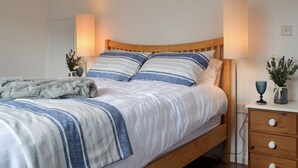 Una cama bien hecha con ropa de cama a rayas, un cabecero de madera, lámparas de noche, una mesita de noche con un florero y un enchufe de pared.