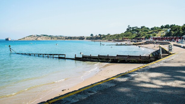 Una playa costera con un muelle que se adentra en el agua, una orilla arenosa y colinas al fondo.