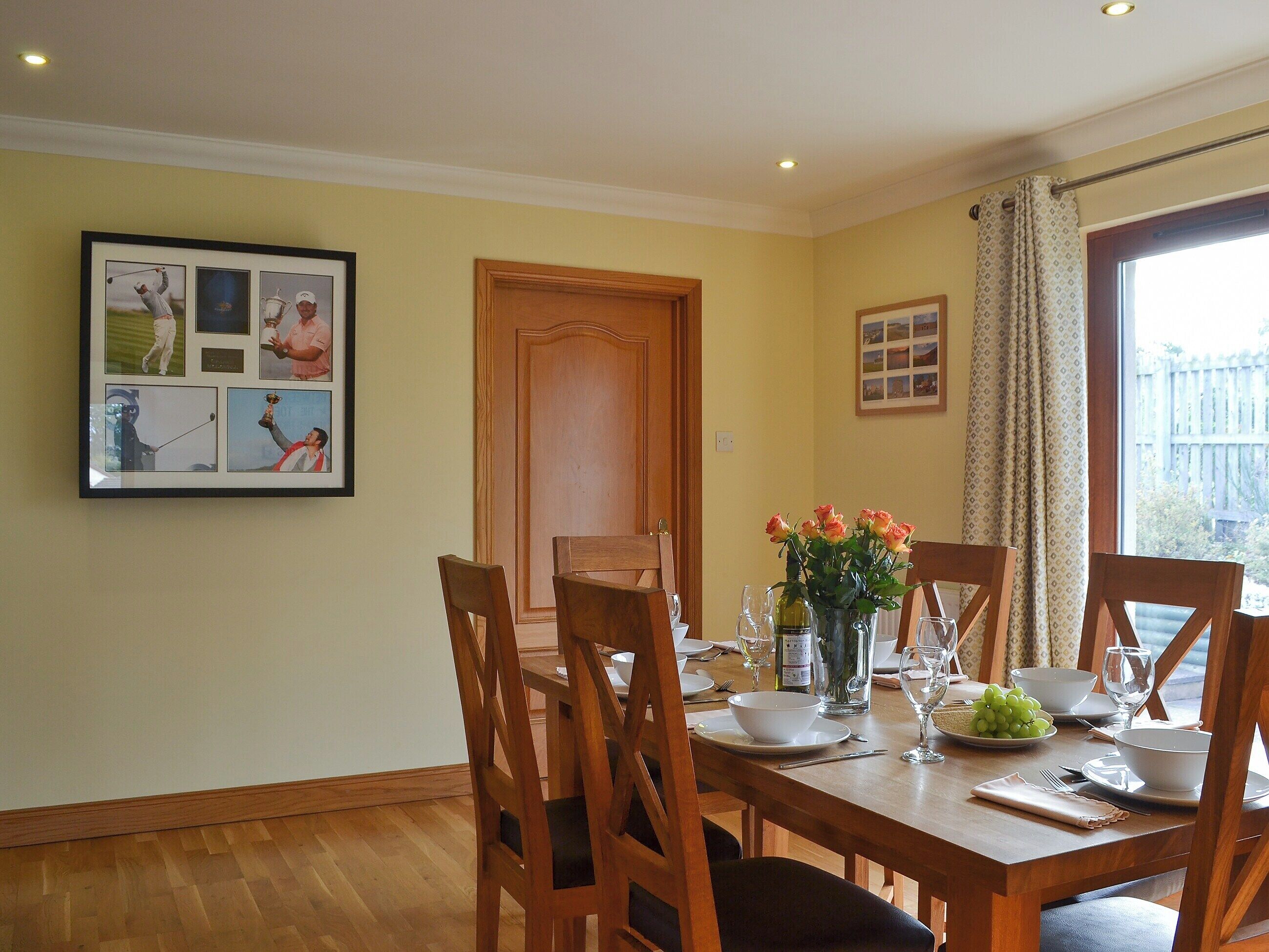 Een eetkamer met een houten tafel voor vier personen, omringd door stoelen. Aan de muur hangt een lijst met een collage van golfafbeeldingen, een houten deur en een raam met gordijnen.