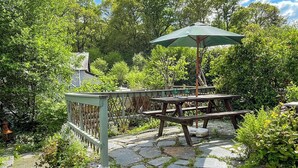 Une terrasse extérieure avec une table en bois et des bancs, entourée d’une végétation luxuriante et de sentiers en pierre.