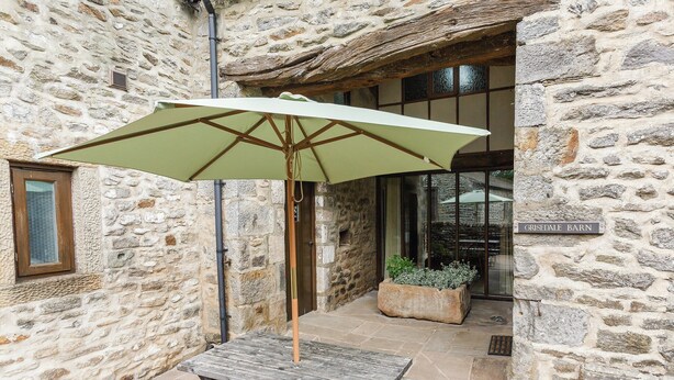 A stone building with a wooden table and bench under a green umbrella, a sign reading "GRISEDALE BARN", and a planter with plants.