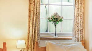 Un dormitorio con mesita de noche de madera, lámpara, cama con ropa blanca y ventana con cortinas florales y un jarrón de flores.