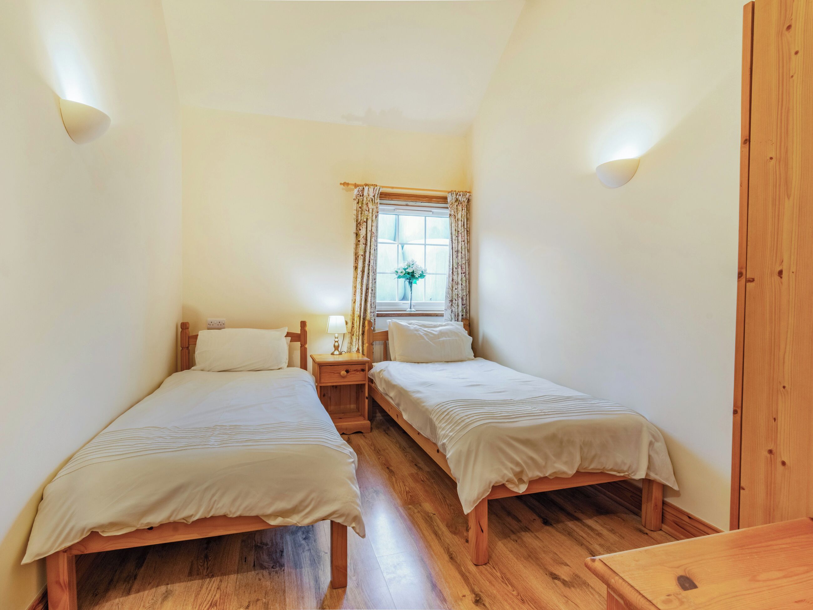 A room with two wooden beds, a small bedside table with a lamp, a window with curtains, and a wooden wardrobe.