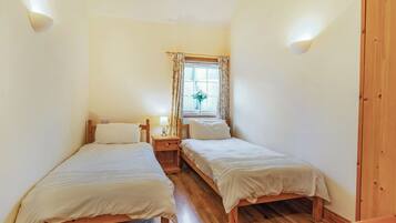 A room with two wooden beds, a small bedside table with a lamp, a window with curtains, and a wooden wardrobe.