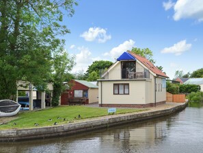 A small house with a balcony, a grassy area, and a body of water. - 2 bedroom accommodation in Stalham (Stalham)