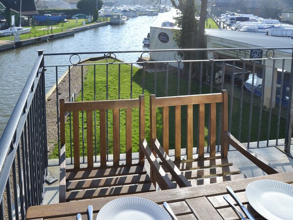 Un balcón con un banco de madera, una mesa con un plato y cubiertos, y vista a un canal con barcos.