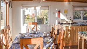 Un comedor con una mesa dispuesta para cuatro, sillas de madera, un ventanal con vistas a zonas verdes, una zona de cocina con hervidor y tostadora, y un extintor de incendios en la pared.