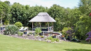 A gazebo with a thatched roof, surrounded by a garden with various plants and a small pond.
