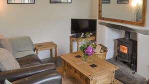A living room with a sofa, a wooden coffee table with a vase of flowers, a television, and a wood-burning stove. - 2 bedroom accommodation in Trewalder, near Wadebridge (Trewalder, near Wadebridge)