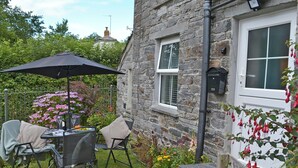 A stone house with a patio area featuring a table and chairs under a black umbrella, surrounded by lush greenery and flowering plants. - 2 bedroom accommodation in Trewalder, near Wadebridge (Trewalder, near Wadebridge)