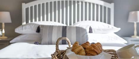 A neatly made bed with white bedding and striped pillows, a wicker basket with croissants, a glass of orange juice, and a nightstand with a lamp.