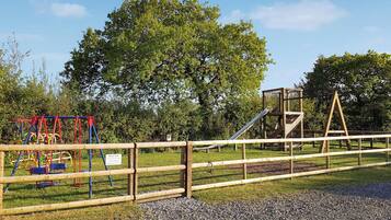 Un parque infantil con columpios, toboganes y estructuras para escalar, rodeado por una cerca de madera y suelo de grava.
