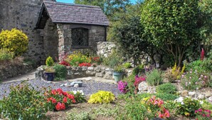 Una cabaña de piedra con techo inclinado, rodeada por un jardín con diversas plantas en flor y un sendero de grava.
