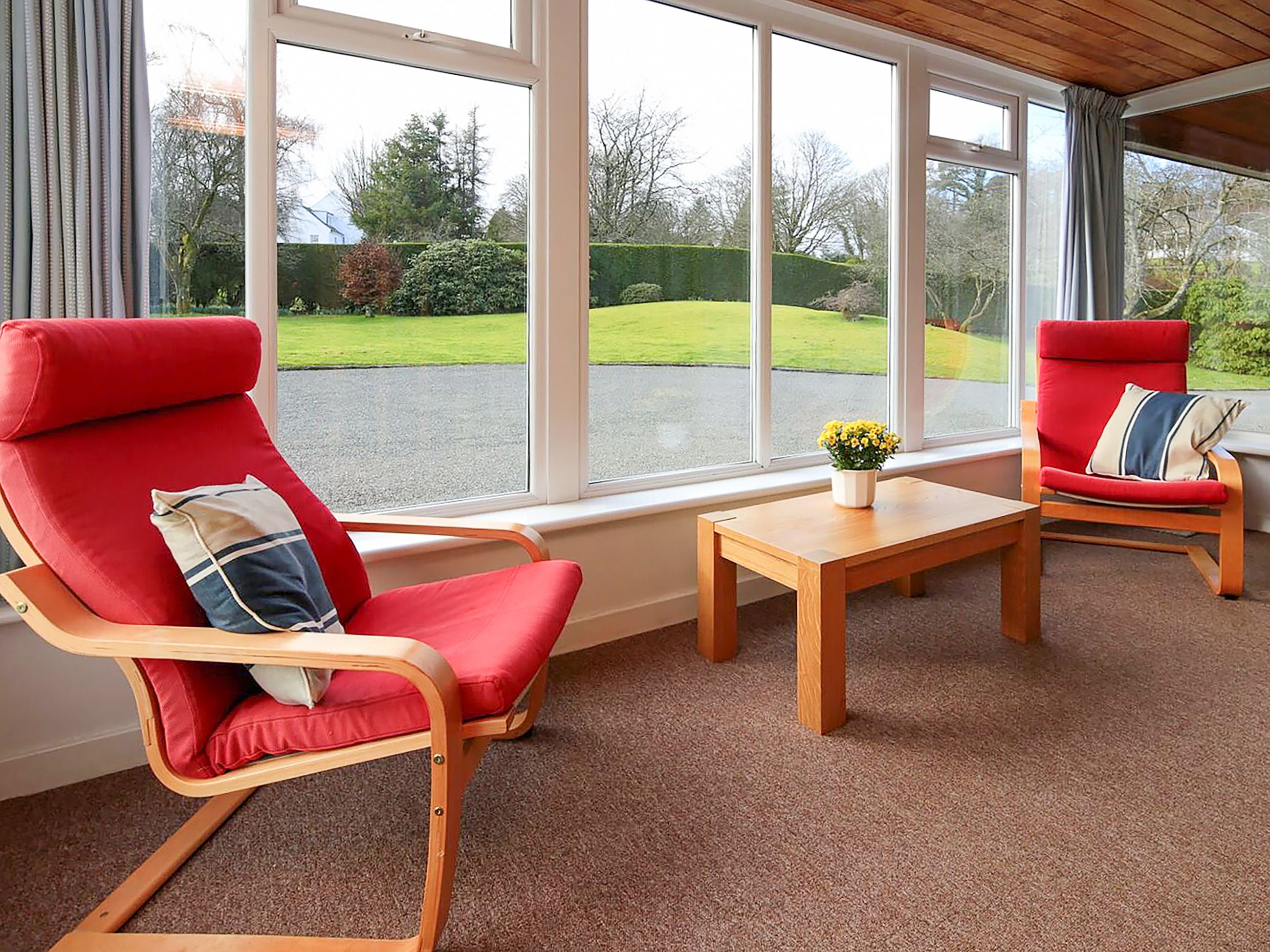 A room with two red chairs, a wooden table, and large windows offering a view of a green lawn and trees.
