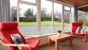A room with two red chairs, a wooden table, and large windows offering a view of a green lawn and trees. - 4 bedroom accommodation in Gatehouse of Fleet, Kirkcudbright (Gatehouse of Fleet, Kirkcudbright)