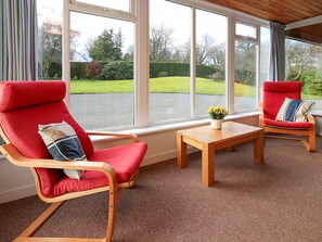 A room with two red chairs, a wooden table, and large windows offering a view of a green lawn and trees. - 4 bedroom accommodation in Gatehouse of Fleet, Kirkcudbright (Gatehouse of Fleet, Kirkcudbright)