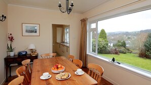 A dining room with a wooden table set for four, a view of the garden through a large window, and a kitchen visible through an open door. - 4 bedroom accommodation in Gatehouse of Fleet, Kirkcudbright (Gatehouse of Fleet, Kirkcudbright)