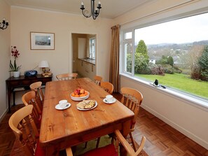 A dining room with a wooden table set for four, a view of the garden through a large window, and a kitchen visible through an open door. - 4 bedroom accommodation in Gatehouse of Fleet, Kirkcudbright (Gatehouse of Fleet, Kirkcudbright)