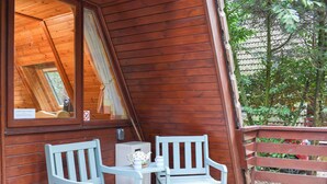 Une cabane en bois dotée d’un balcon, de deux chaises blanches, d’une petite table et d’une fenêtre donnant sur la forêt environnante.