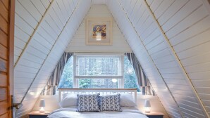Une chambre avec un plafond en pente, une fenêtre avec des stores, un lit avec des oreillers, deux lampes de chevet et un tableau encadré au-dessus du lit.