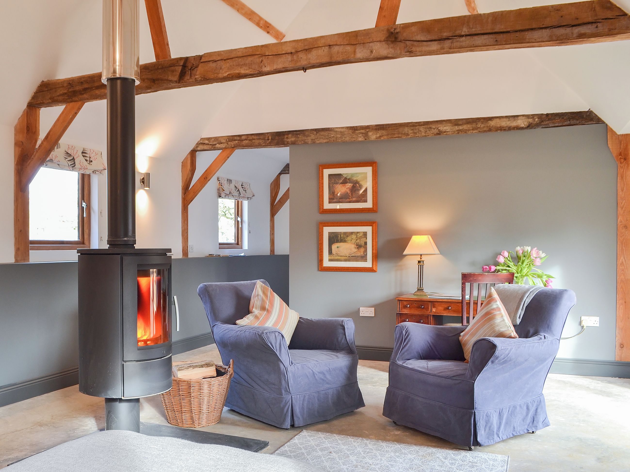 A room with a wood-burning stove, two blue armchairs, a wicker basket, a lamp, and two framed pictures on the wall.