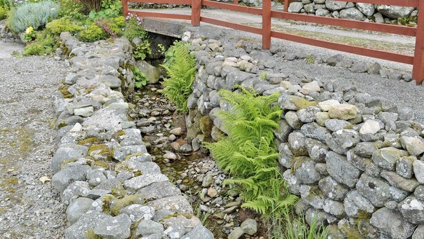 Un puente de piedra sobre un arroyo, rodeado de follaje y una cerca de madera.