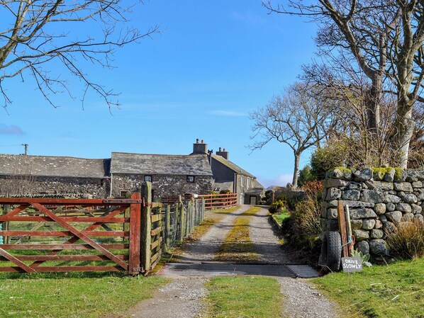 A rural road leading to a stone house with a metal gate and a 'Drive Slowly' sign.
