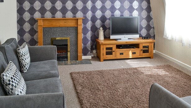 A living room with a fireplace, a television on a wooden stand, and a grey sofa with patterned cushions. - 3 bedroom accommodation in Cromer (Cromer)