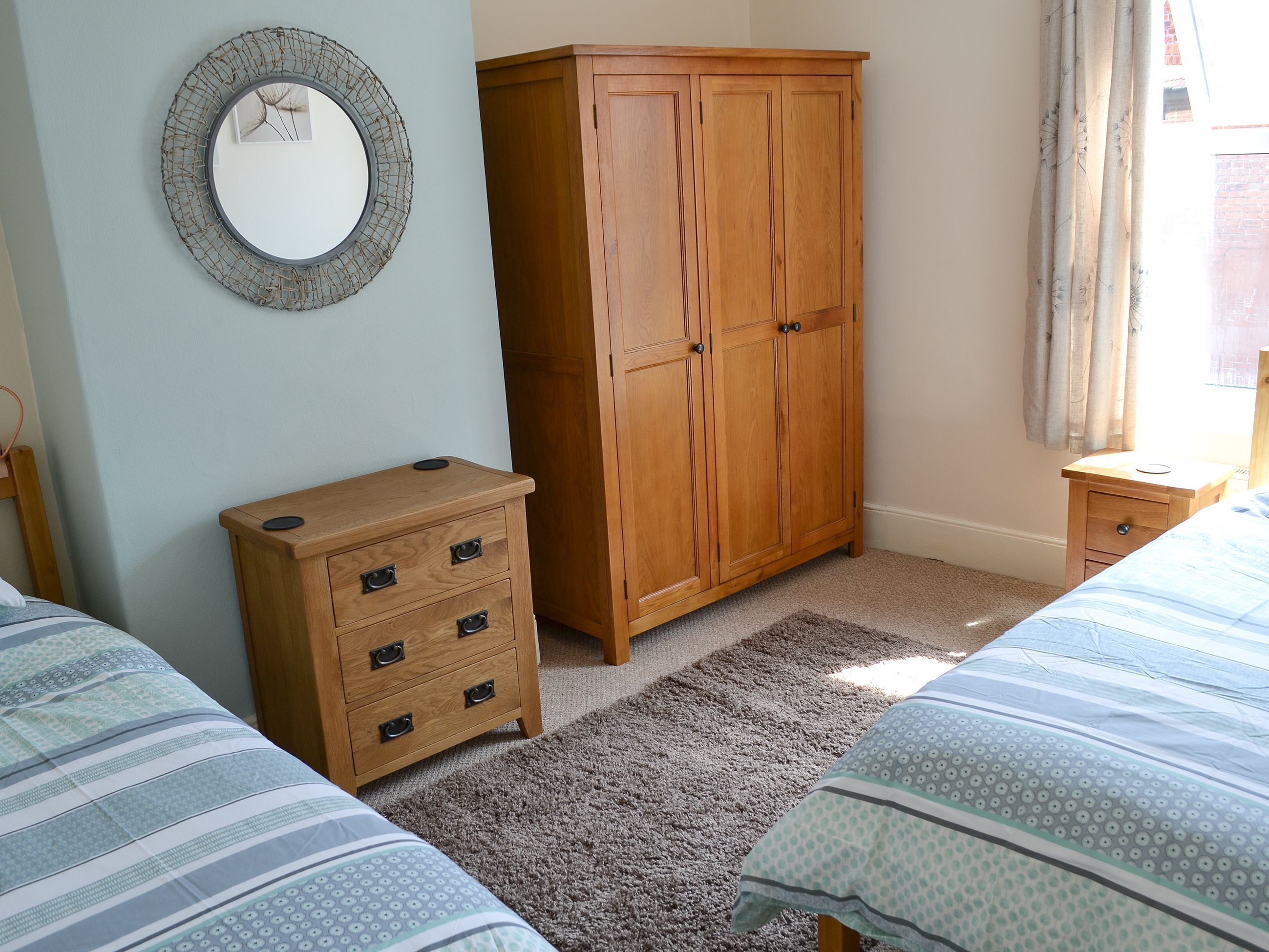 Une chambre avec deux lits, une armoire en bois, une petite commode, un miroir rond et une fenêtre avec des rideaux.