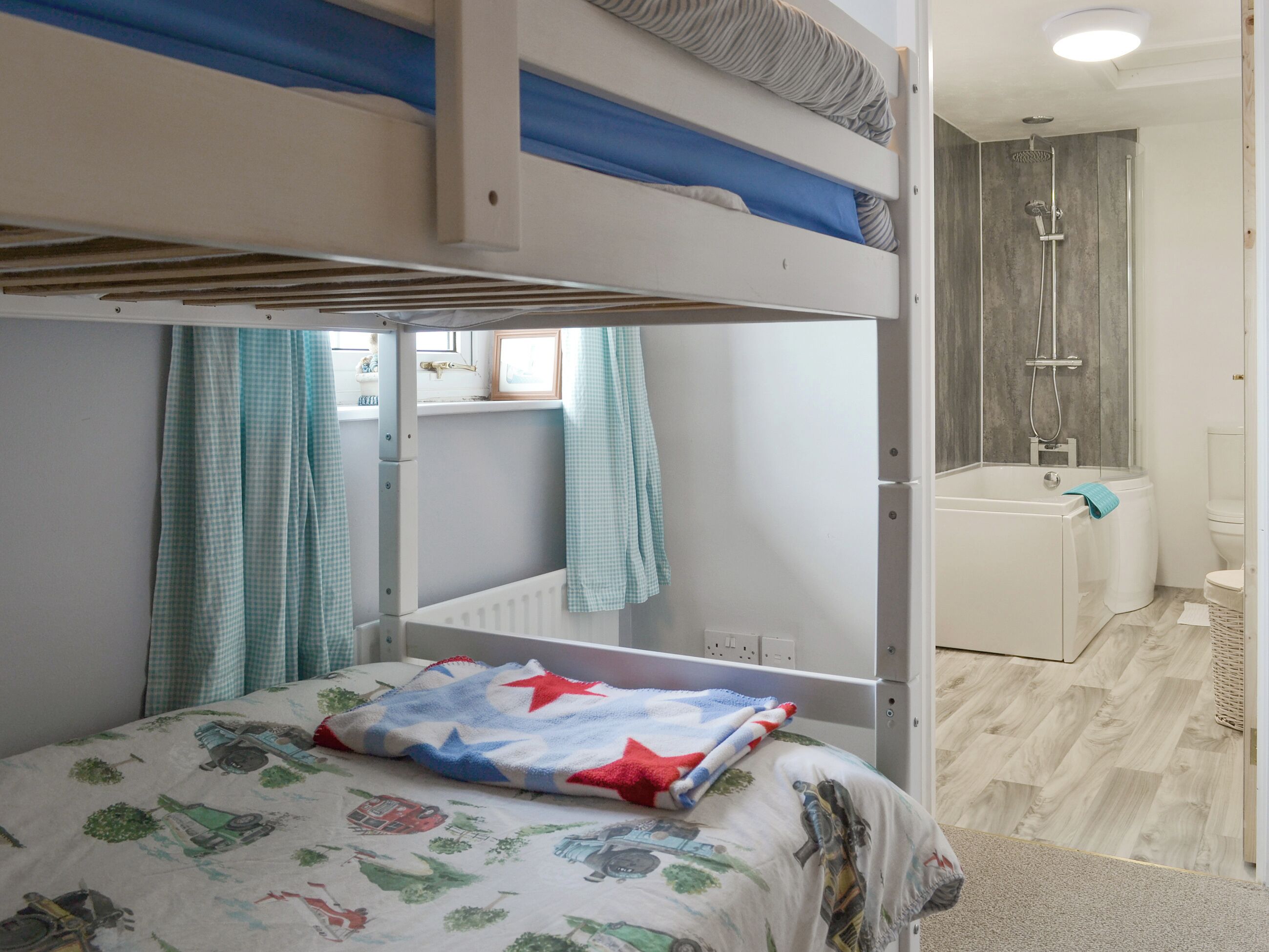A bunk bed with a checkered curtain, a bed with a vehicle-themed blanket, a bathroom with a shower, and a toilet.