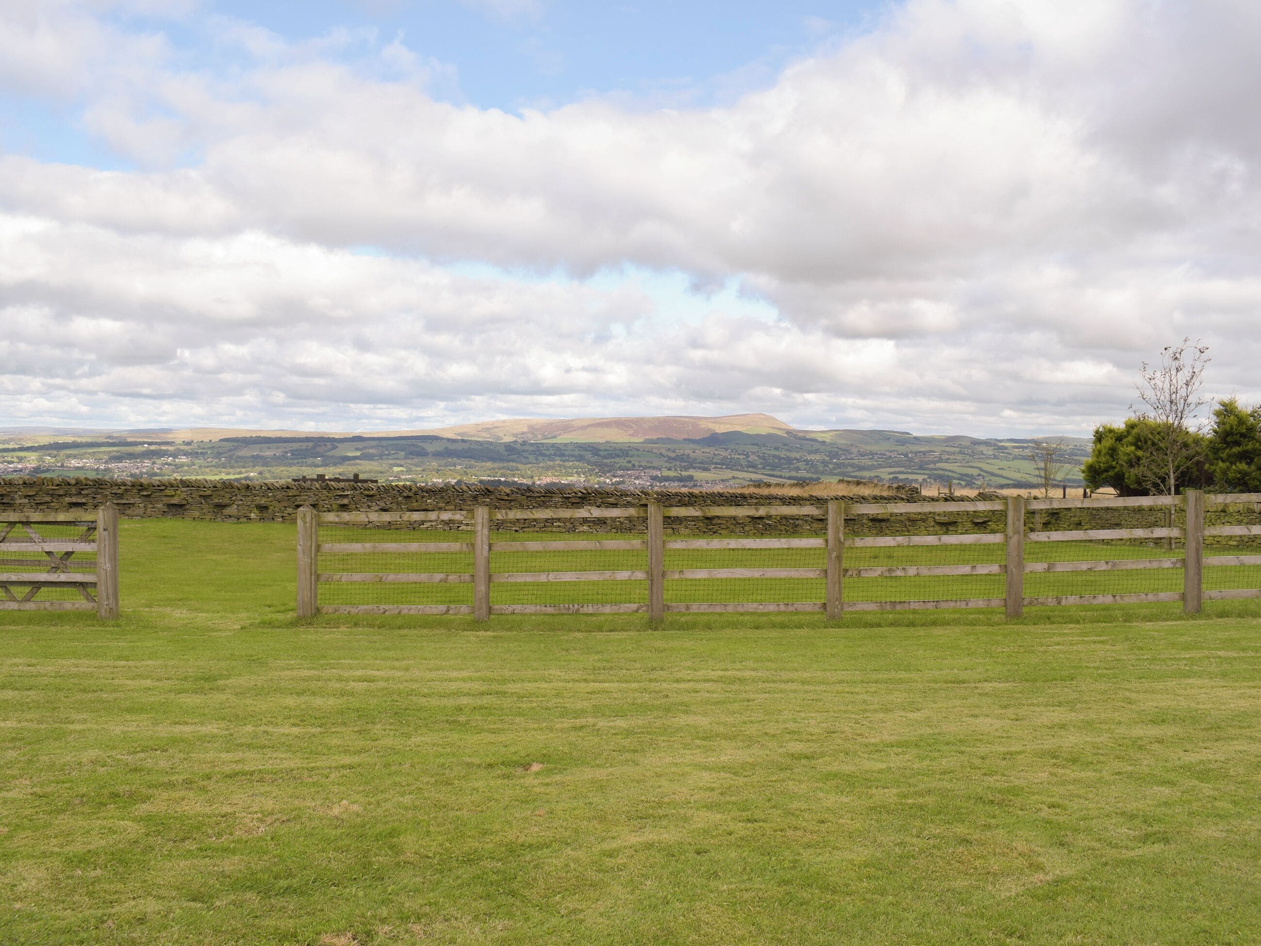 A rural landscape with a stone wall, wooden fence, and a view of distant hills and a small town.
