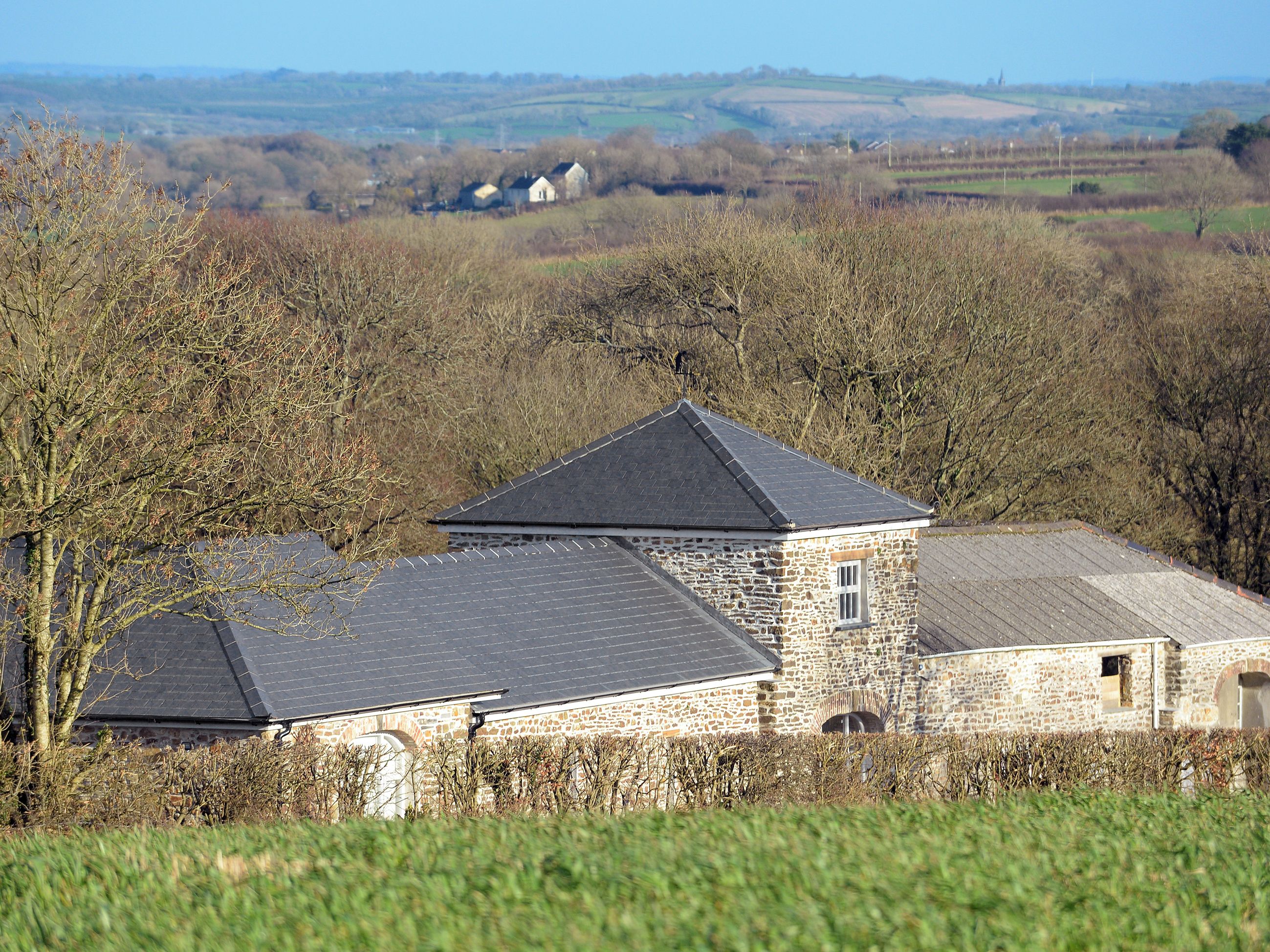 A stone building with a sloped roof, surrounded by trees and fields.
