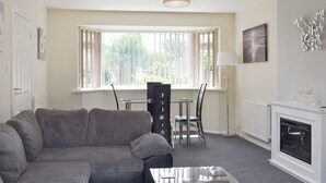A living room with a sofa, a dining table with chairs, a fireplace, and a large window with blinds. - 1 bedroom accommodation in Broadstone, near Bournemouth (Broadstone, near Bournemouth)