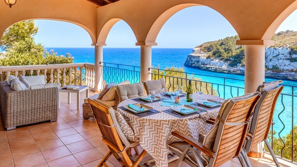 A covered outdoor seating area with a table set for dining, overlooking a coastal view. - Fantastic Villa "Calima" with Sea Views, Loggia, Air Conditioning, and Wi-Fi (S'Estany den Más)