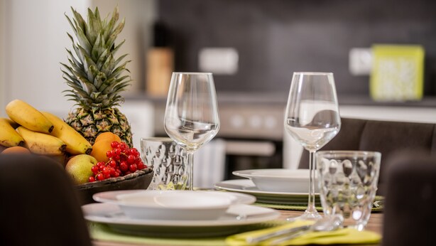 A dining table set with plates, glasses, and a fruit basket containing bananas, a pineapple, and red berries.