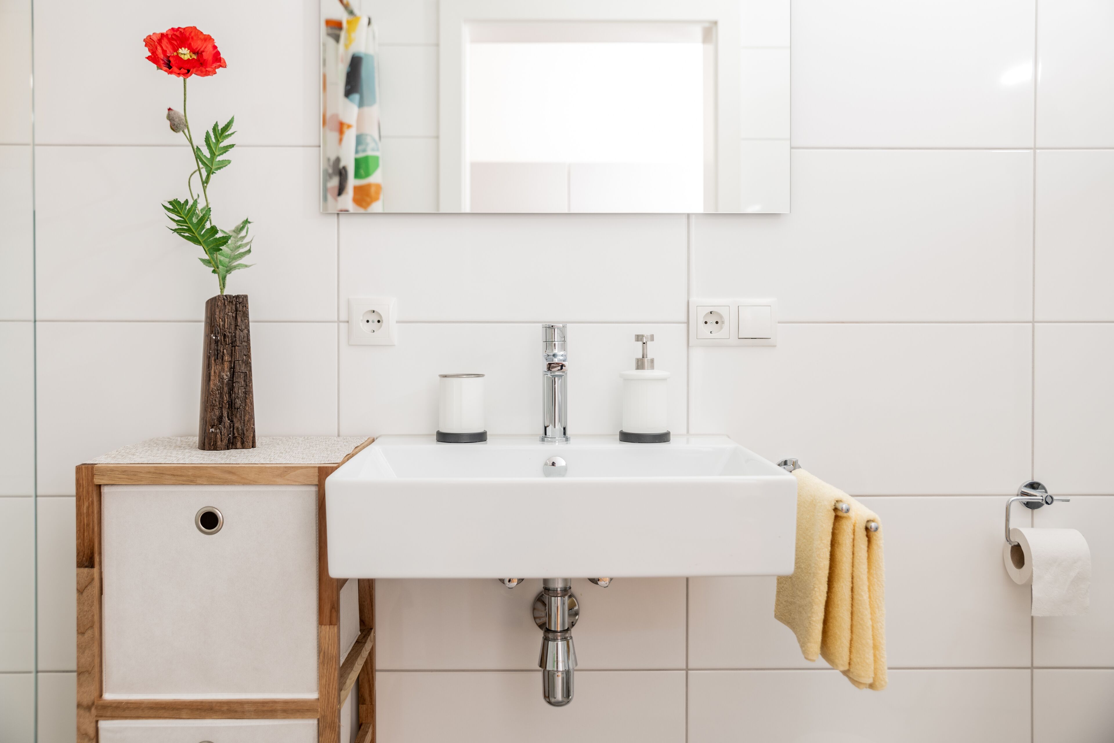 A bathroom with a white sink, a mirror, a wooden cabinet, a red flower in a vase, and a yellow towel hanging on the wall.