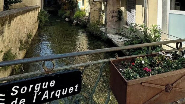 Ein schmaler Kanal, der von Gebäuden mit Balkonen gesäumt wird, mit einem Schild, auf dem 'Sorgue de l'Arquet' steht.