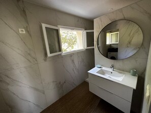 Un baño con pared de mármol, lavamanos blanco, espejo y ventana.